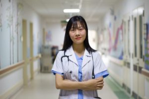 Une soignante en blouse se tient dans un couloir d'hôpital, l'air accueillant.